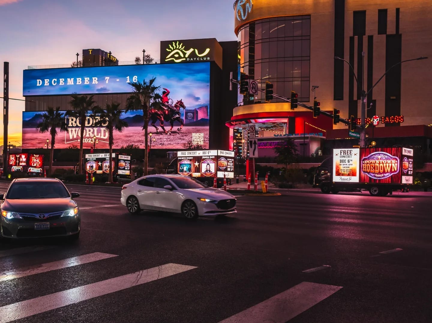 Urban street at twilight with illuminated billboards and cars.