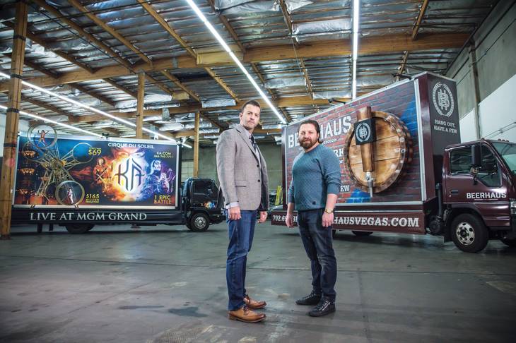 Two men standing in warehouse with branded trucks.