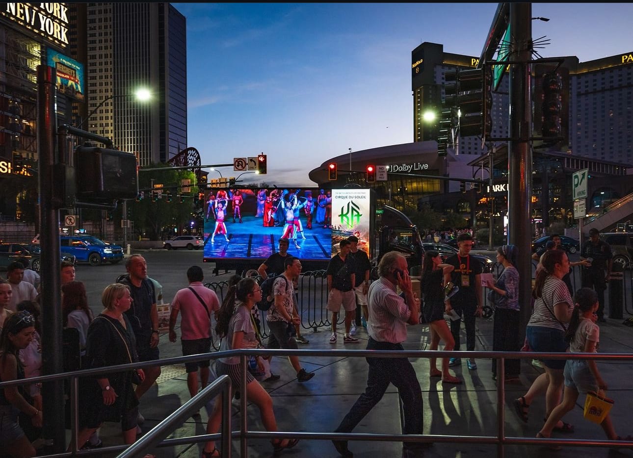 Crowded street corner with large outdoor screen at dusk. Cirque du Soleil